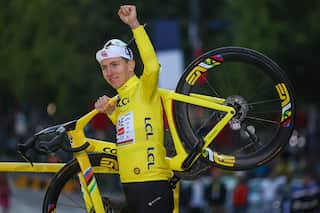 A man wearing yellow cycling jersey and a white cap holds his left arm up in triumph as he carries a yellow bicycle over his right shoulder