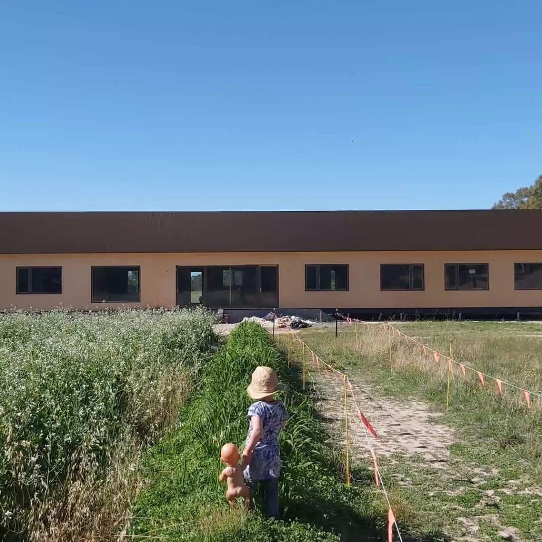 A toddler in a bucket hat holds a doll by the hand, facing away towards a long building with windows