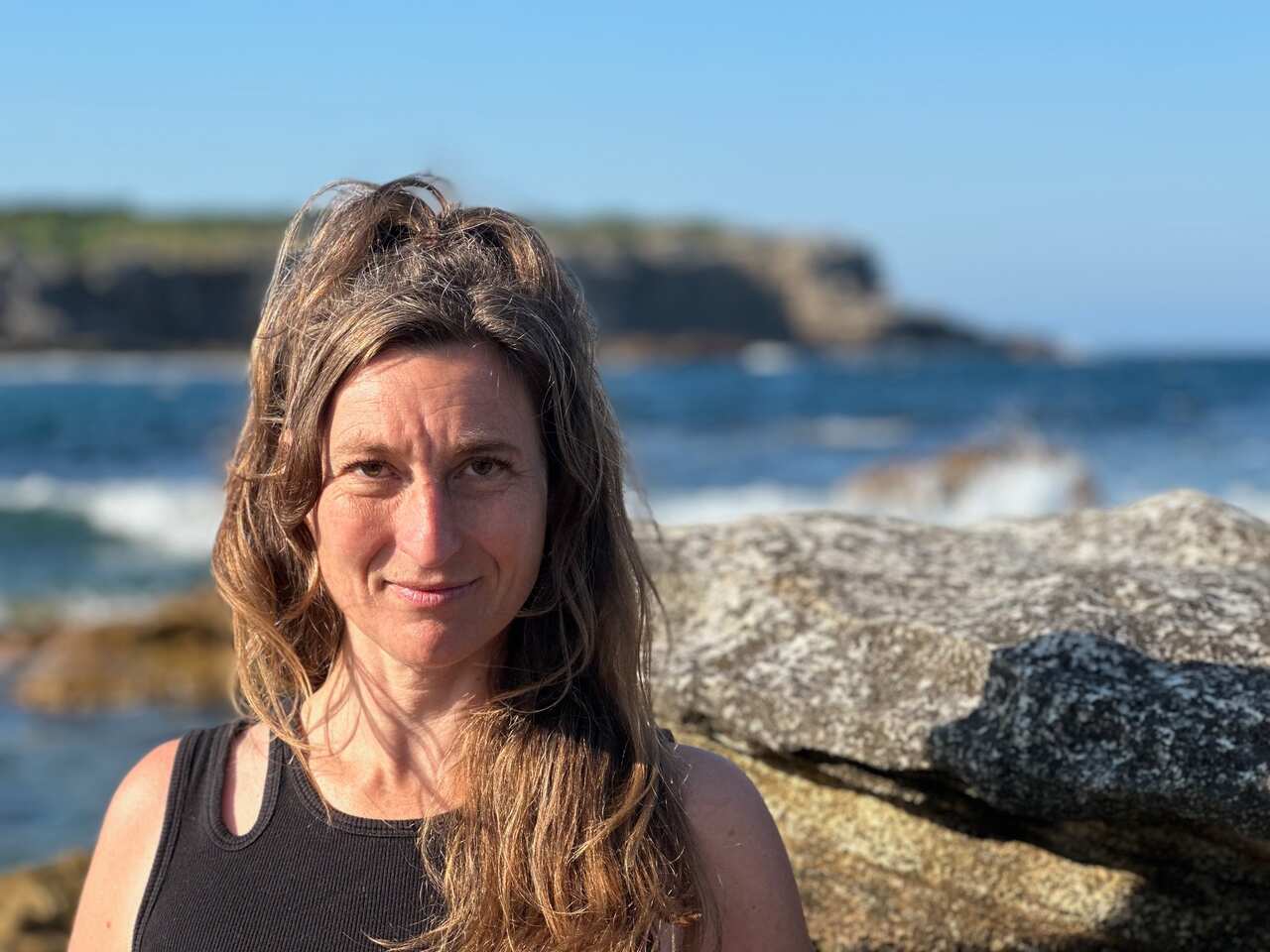 A woman in a black tank top and with long brown hair looks at the camera with a neutral expression. The ocean is in the background.