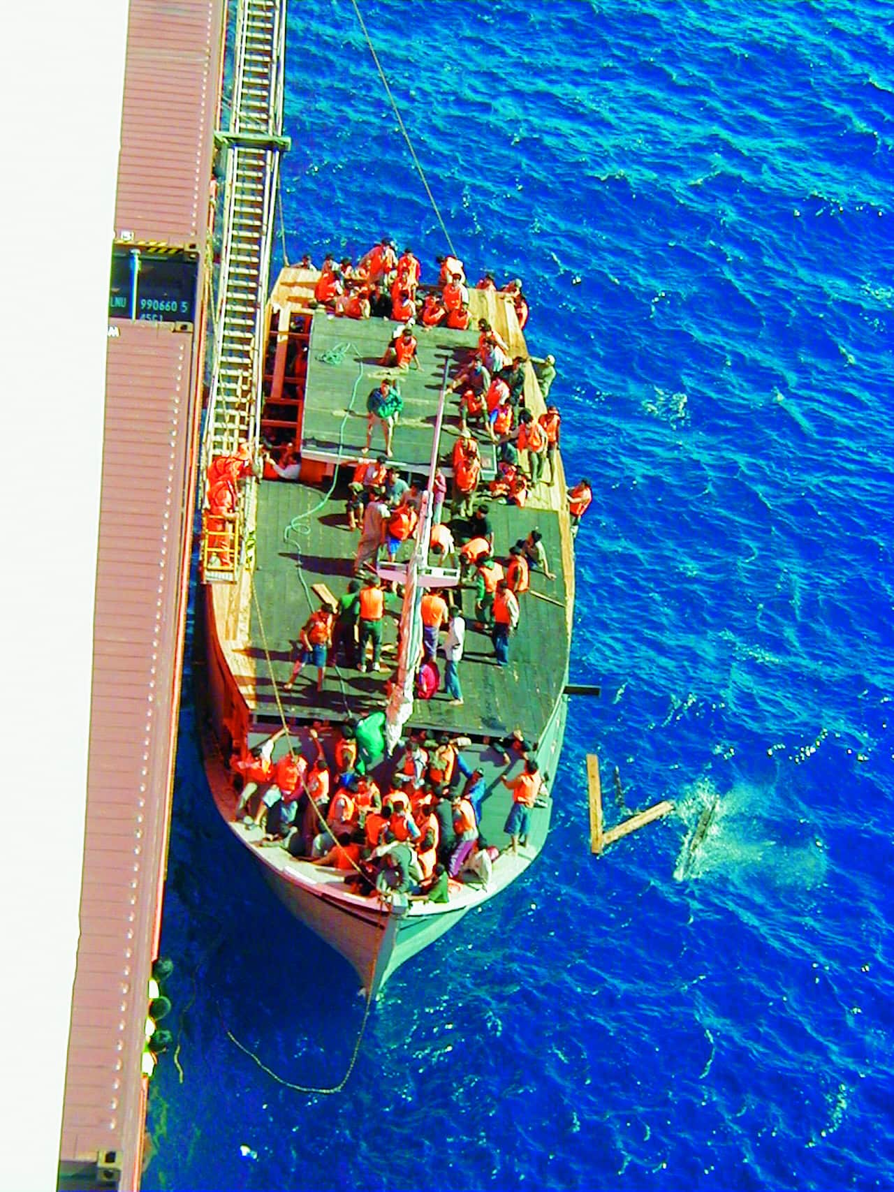 A small boat loaded with people next to a big cargo ship in blue waters.