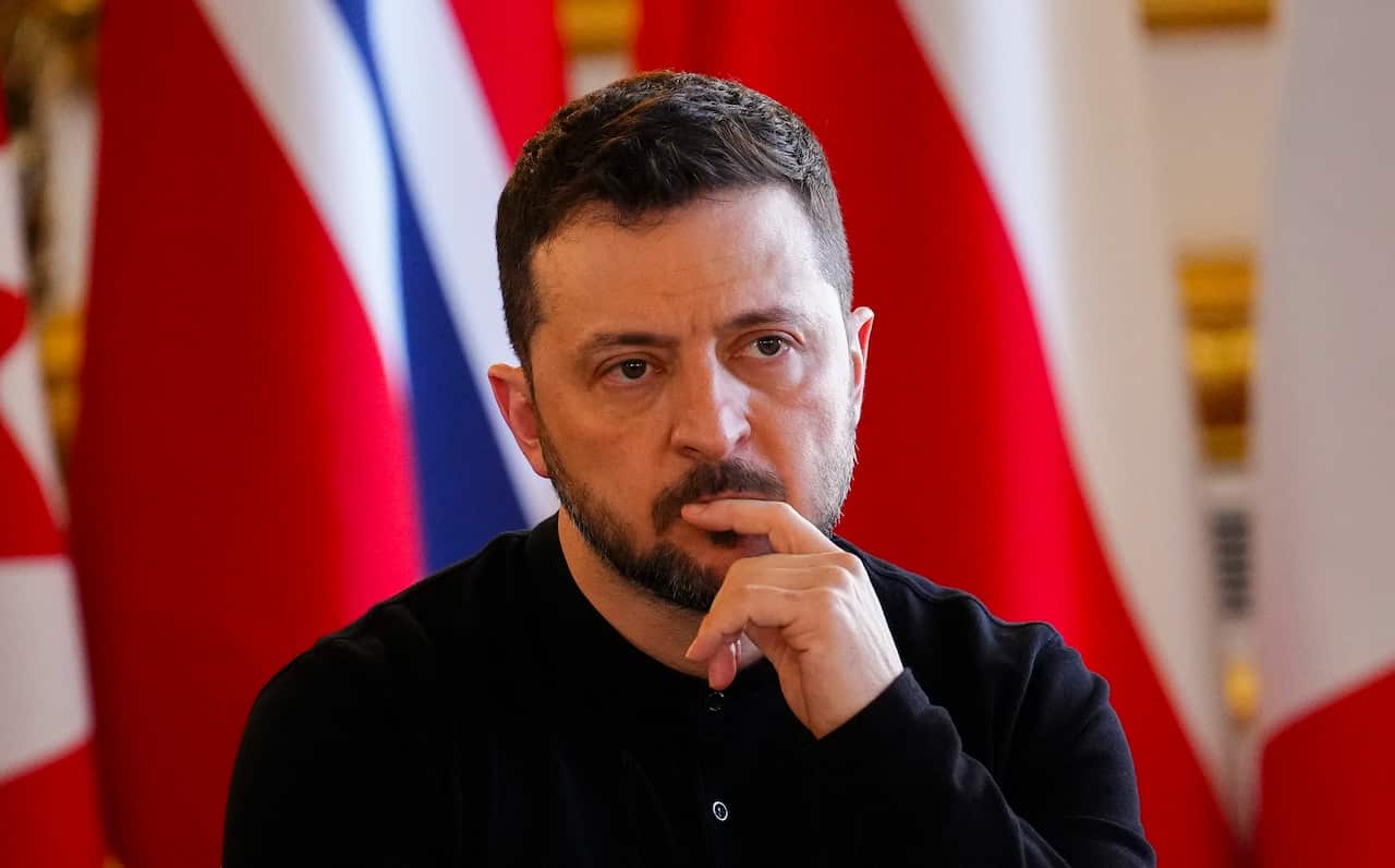 A man in a black crew neck shirt sits, with his finger resting on his lips in a thoughtful pose.