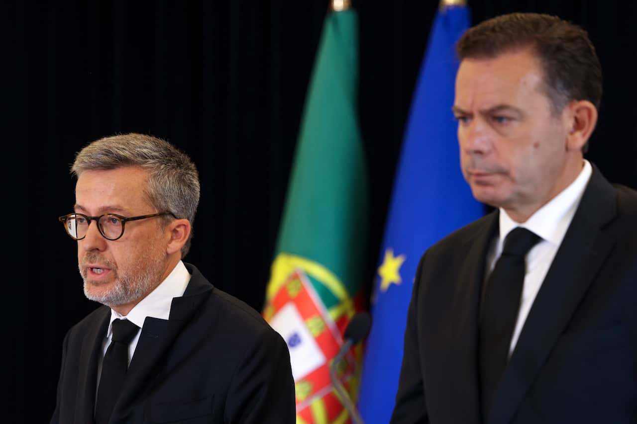 Two men in black speak to the media as they stand in front of flags.