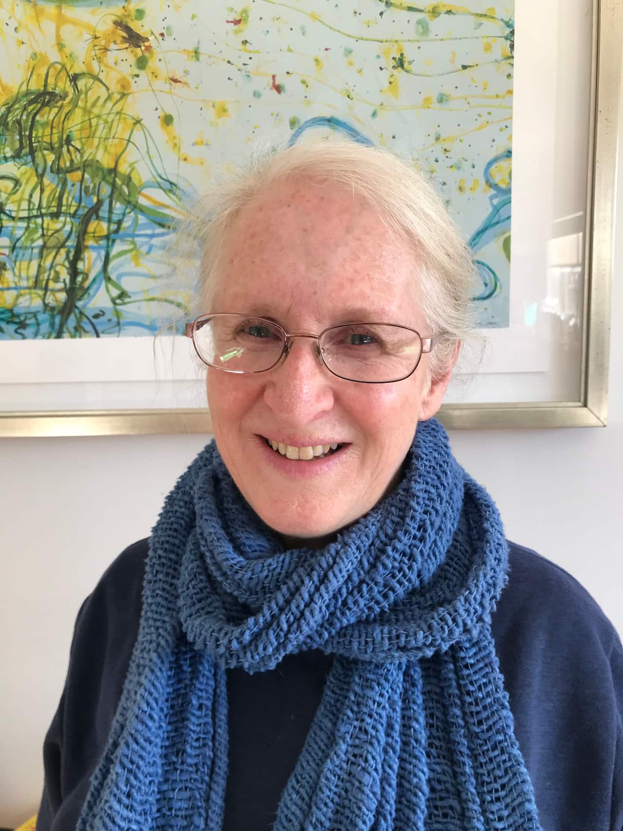 A lady wearing a knitted blue scarf and glasses smiles.