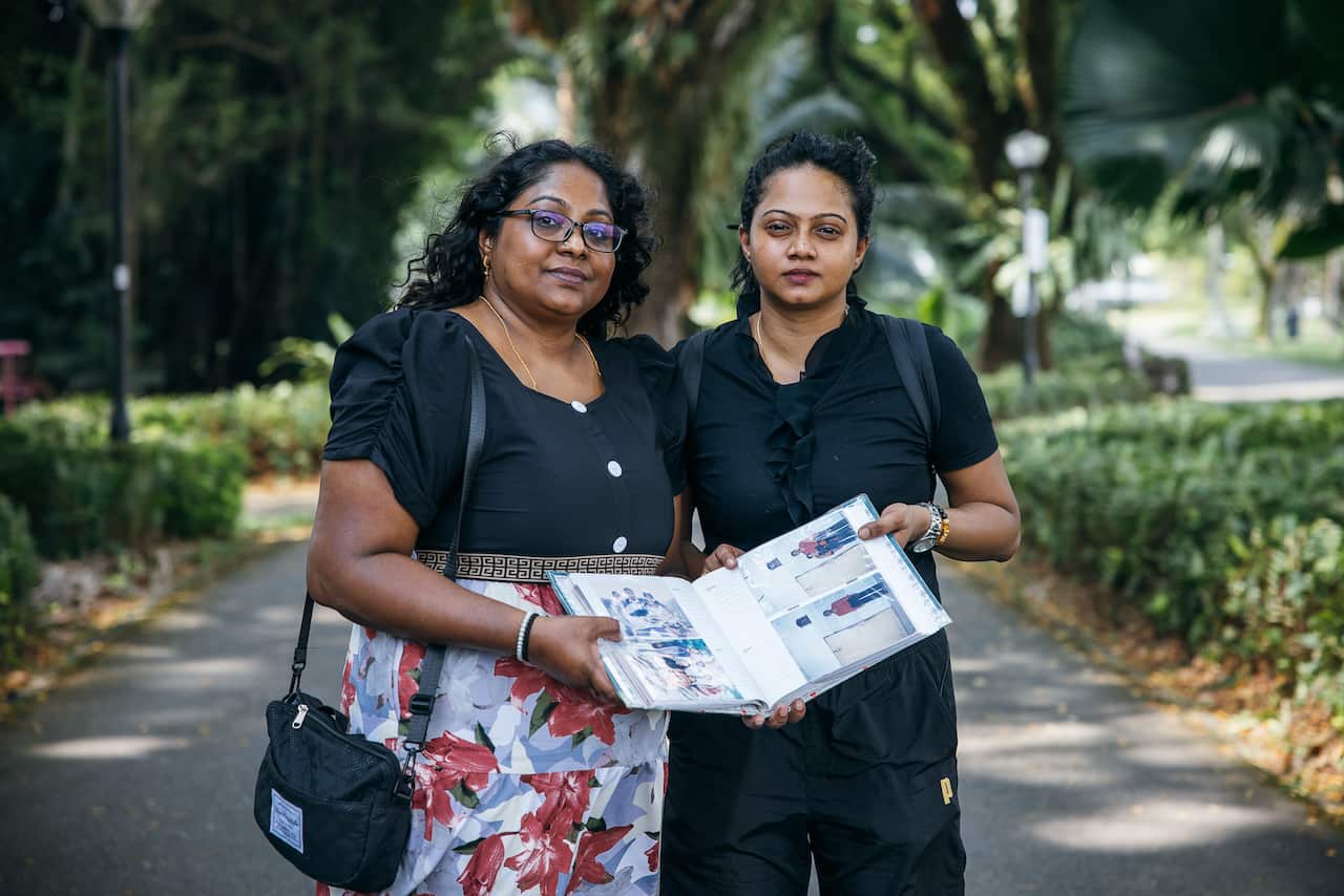 Two women stand on a path in a park, holding open a photo album. They both have neutral facial expressions
