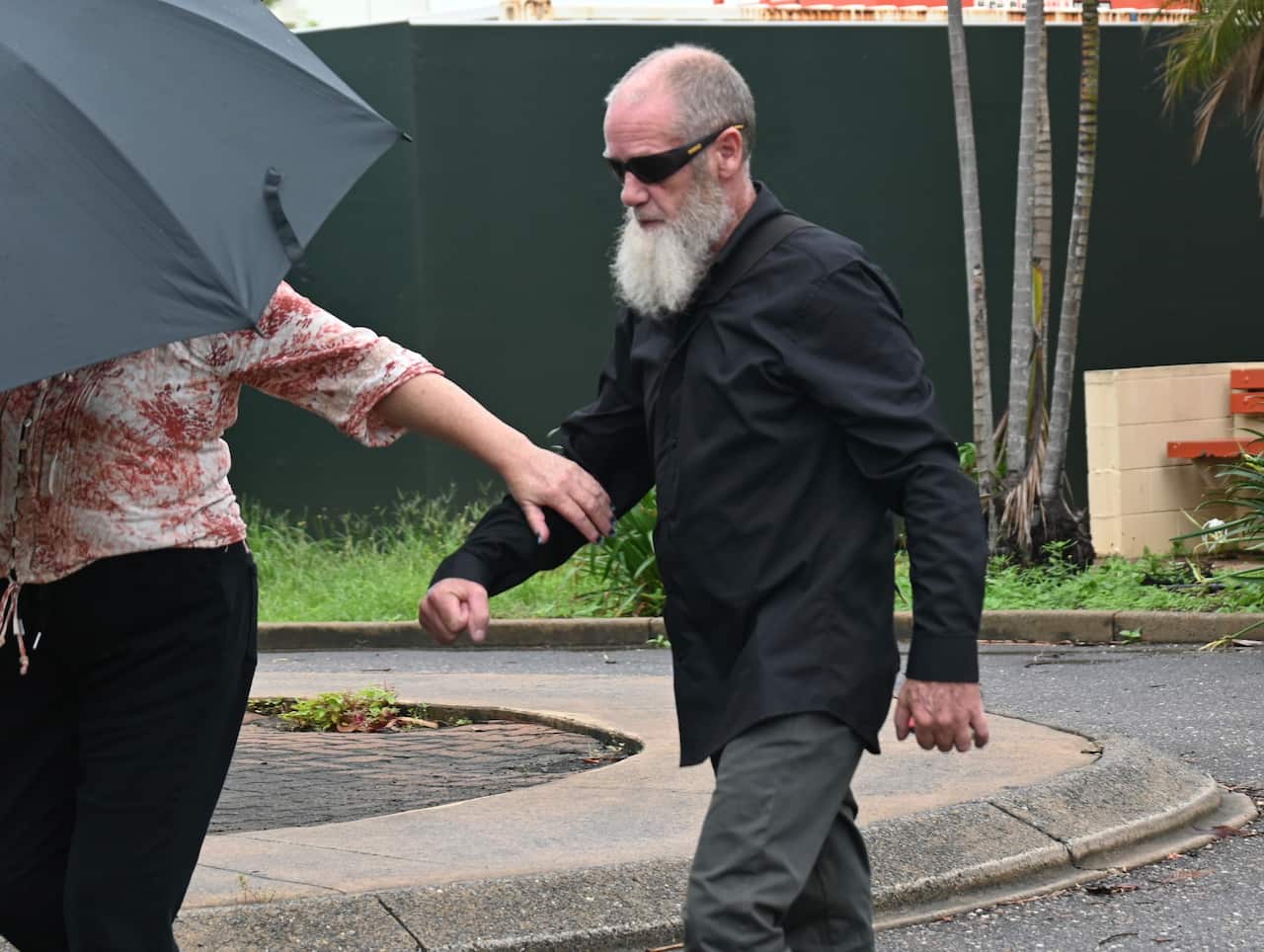 A man with a white goatee beard and wearing sunglasses walking outside. He's wearing a long-sleeved black shirt.