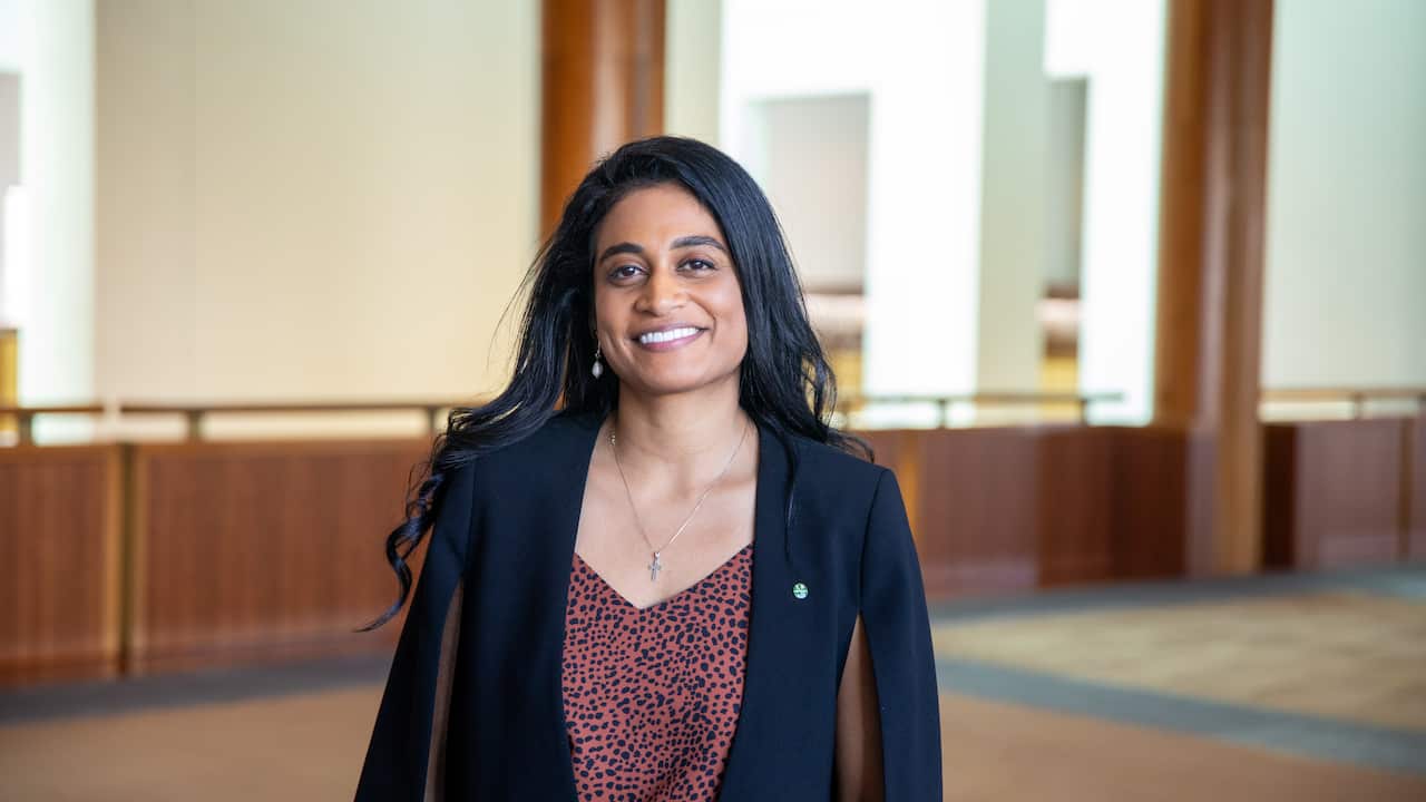 A woman smiling in a parliamentary hallway, wearing a spotted blouse with a black blazer.