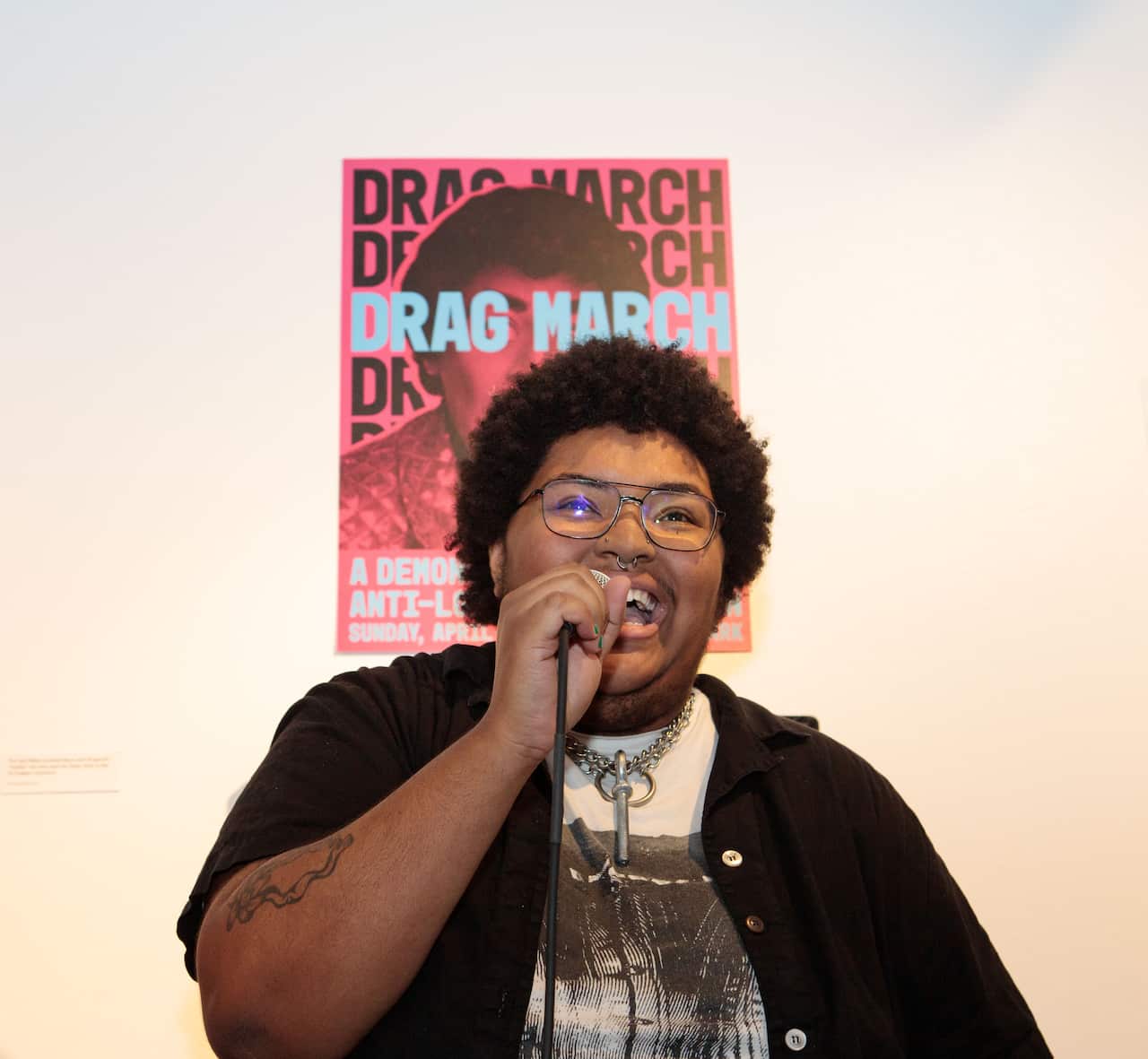 A person wearing a black cardigan over a white round-neck T-shirt smiles as they hold a microphone in front of a wall with a poster.