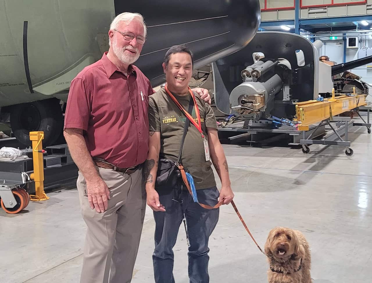 A man in a maroon polo shirt with his arm around the shoulder of a man in an olive green t-shirt who has a dog on a lead next to him. They are standing in front of a RAAF plane in a museum.