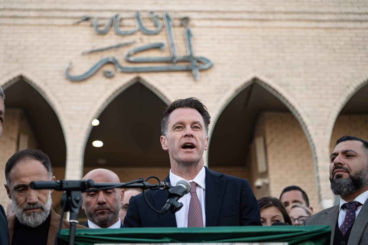 Chris Minns, wearing a suit, speaking into a microphone at a lectern outside a building with Arabic writing on it. There are people behind him