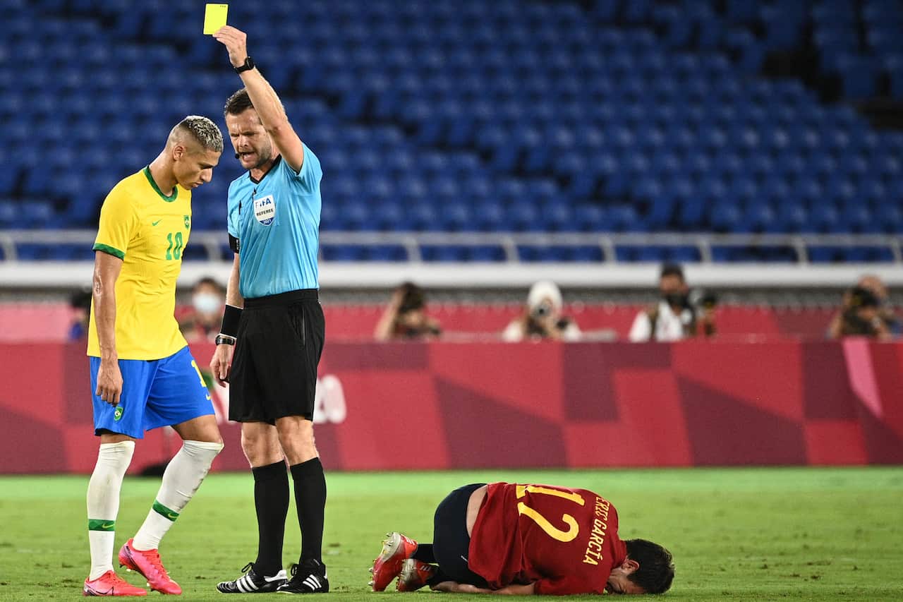 A referee showing a yellow card to a player while another player lies on the ground