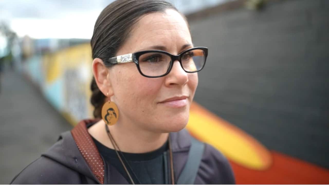 a woman with a braid and black glasses stands in front of a mural of the Aboriginal flag