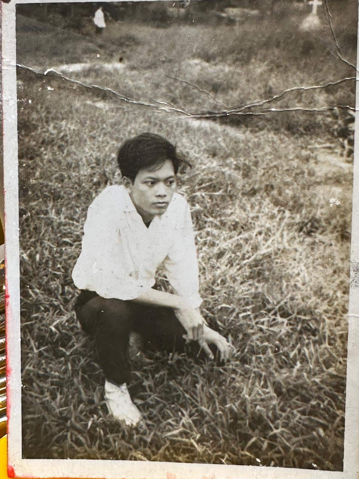 Black and white photo of a young man wearing long dark pants and a long-sleeved white shirt crouching down in a rural setting.