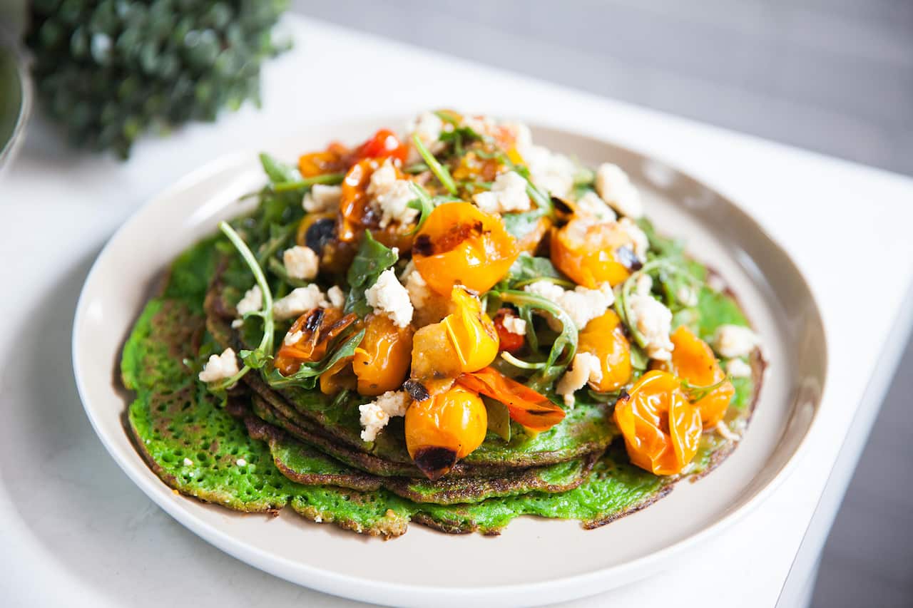 Several vibrant green thin pancakes sit on a plate, topped with a tomato, rocket and feta mixture. 