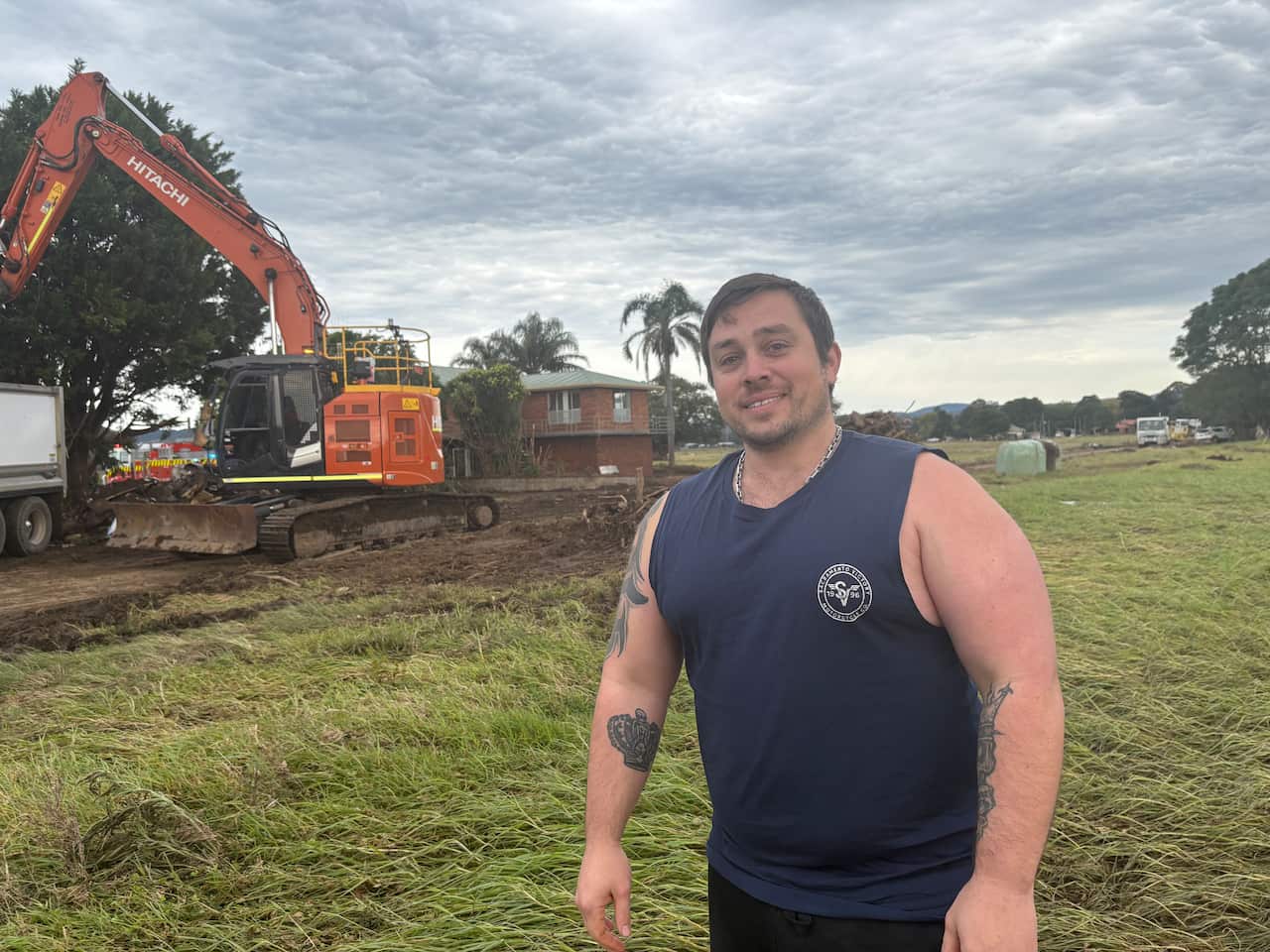 A young, larger set man, with arm tattoos, wearing a dark blue singlet, smiling at the camera. Behind him is a large piece of heavy machinery on a muddy road. The grass appears to be all swept to one side.