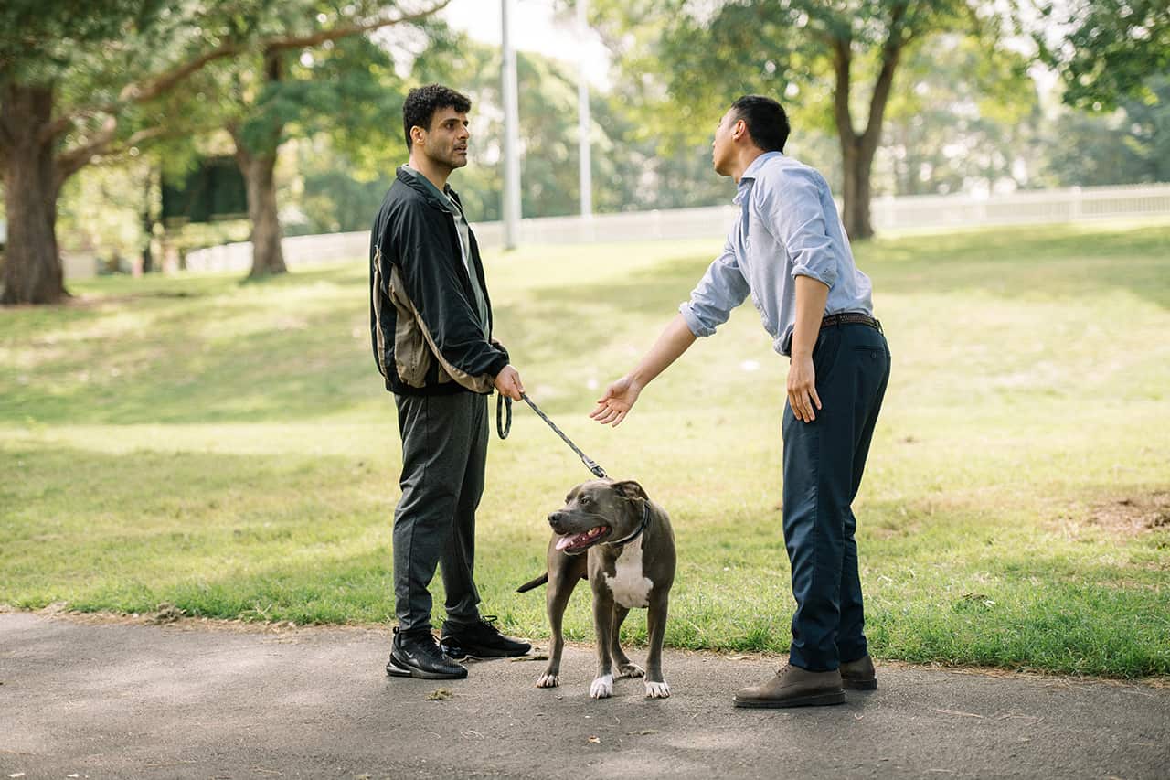 Two men sand on a path in a park. One holds the lead of a dog, the other is reaching towards the lead. 