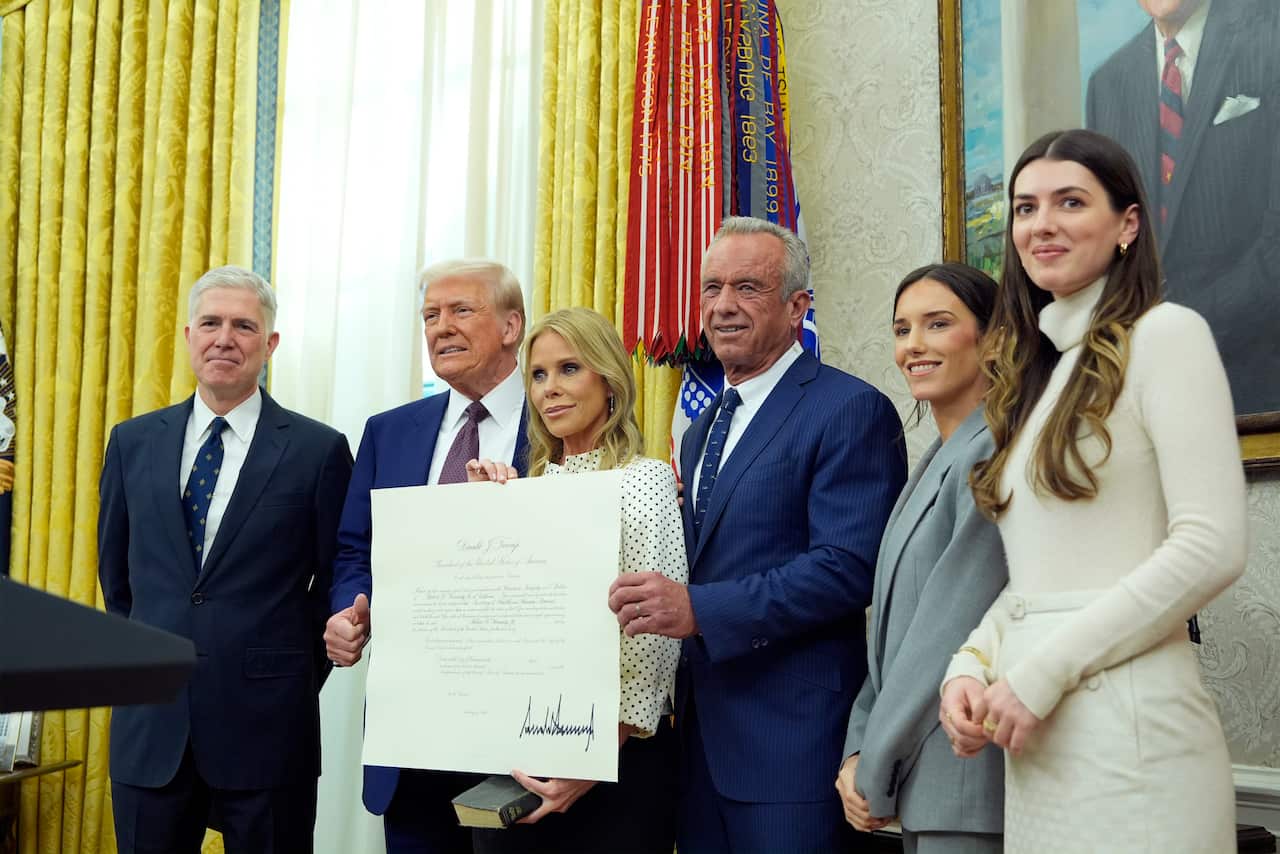 A group of people, including Donald Trump and Robert F Kennedy Jr, standing in the Oval Office. A woman, Cheryl Hines, is holding up a large order.