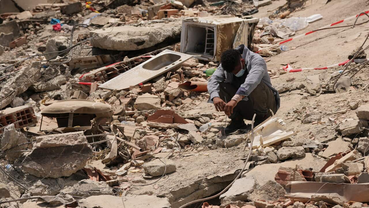 A man in a long-sleeved blue shirt squats amid concrete wreckage. His face is downcast and covered by a surgical mask.