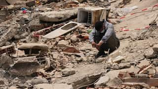 A man in a long-sleeved blue shirt squats amid concrete wreckage. His face is downcast and covered by a surgical mask.