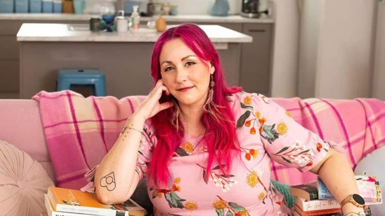 Bigambul and Wakka Wakka author Melanie Saward has bright pink hair and dark brown eyes. She sits on a pink lounge surrounded by stacks of books, looking into the camera.