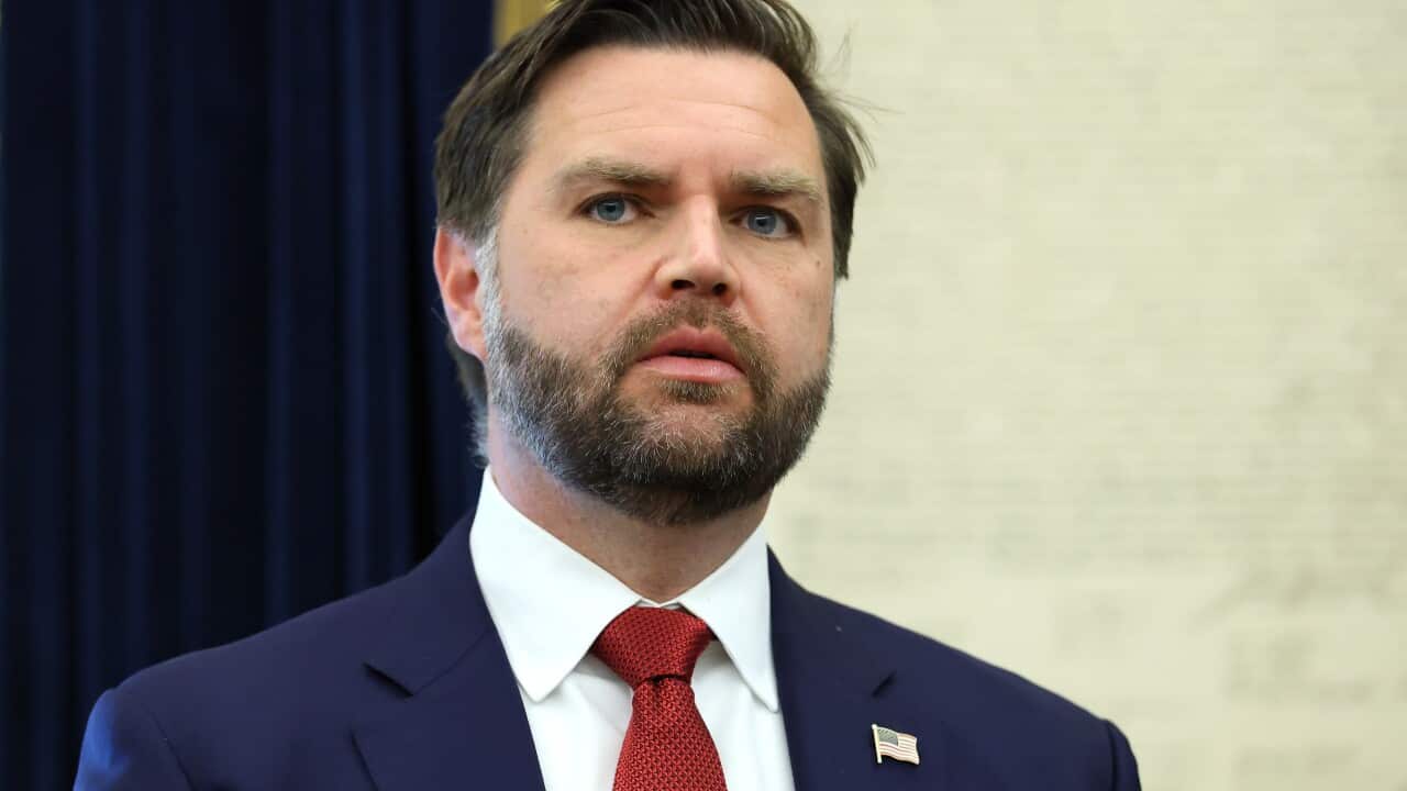 JD Vance, wearing a blue blazer, white shirt, and red tie. The blazer has an American flag pin on it.