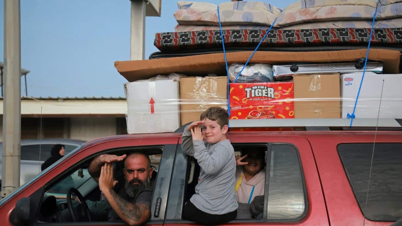 A man and a child gesture from a red car with boxes and mattresses strapped on the roof.