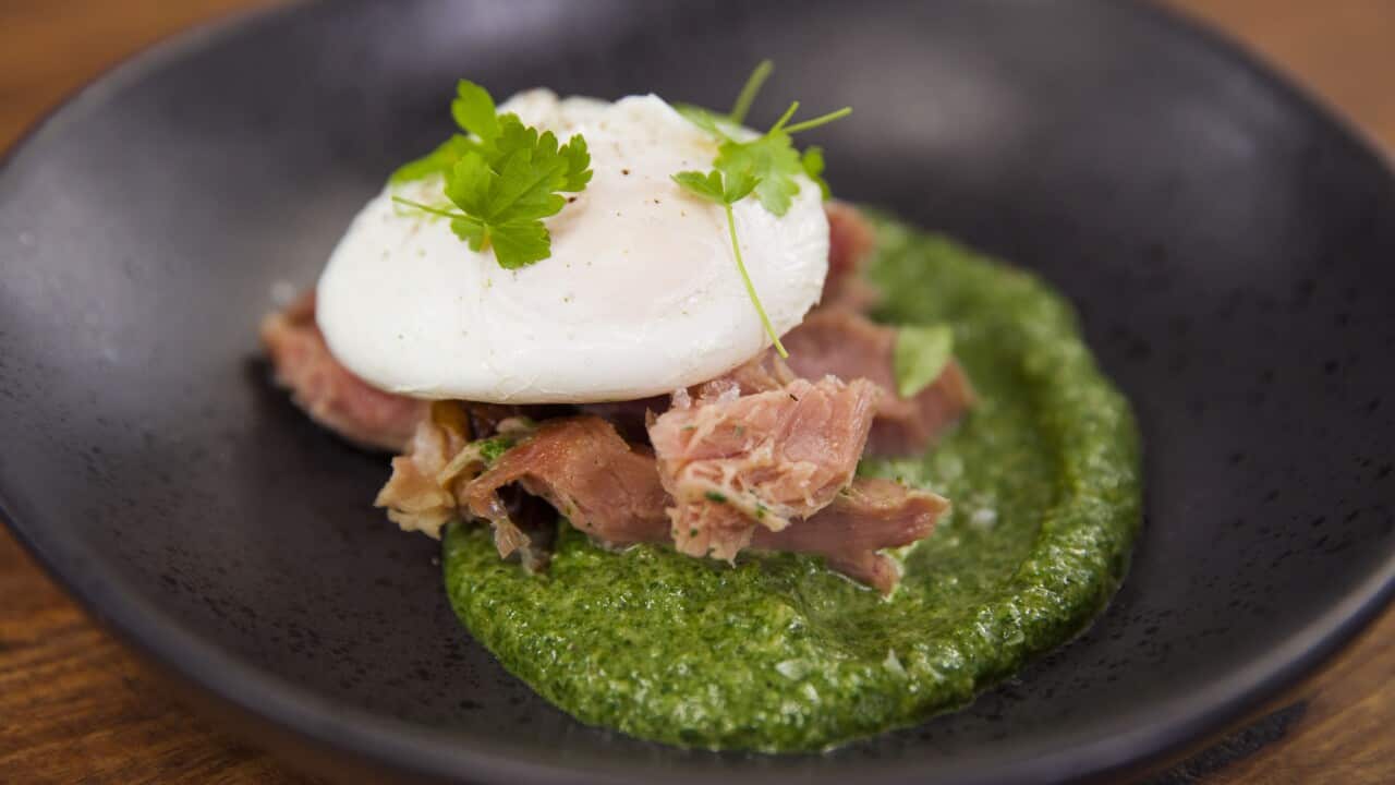 A round of chunky green sauce sits on a wide, dark plate, topped with a pile of cooked ham hock meat and a poached egg.