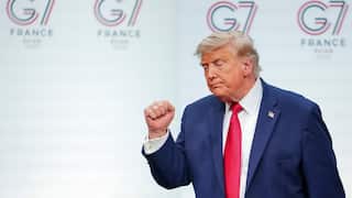 Donald Trump raising a clenched fist while wearing a navy suit and red tie, standing in front of a G7 France backdrop.