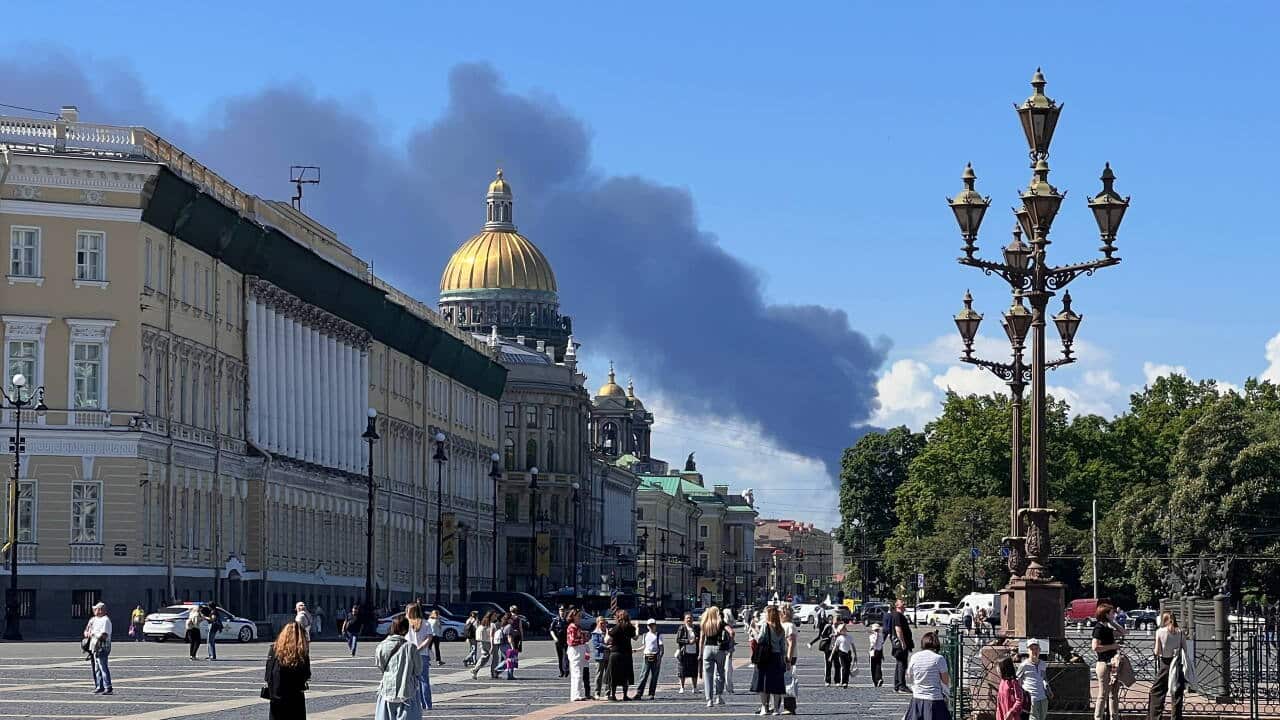 Smoke rising in the distance, in the foreground city buildings and pedestrians walking in a city square