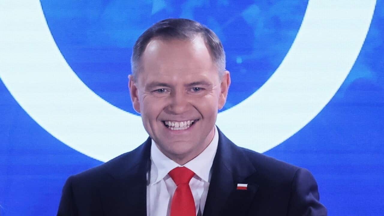 A man in a suit with a Polish flag pin on it, standing in front of a blue background.