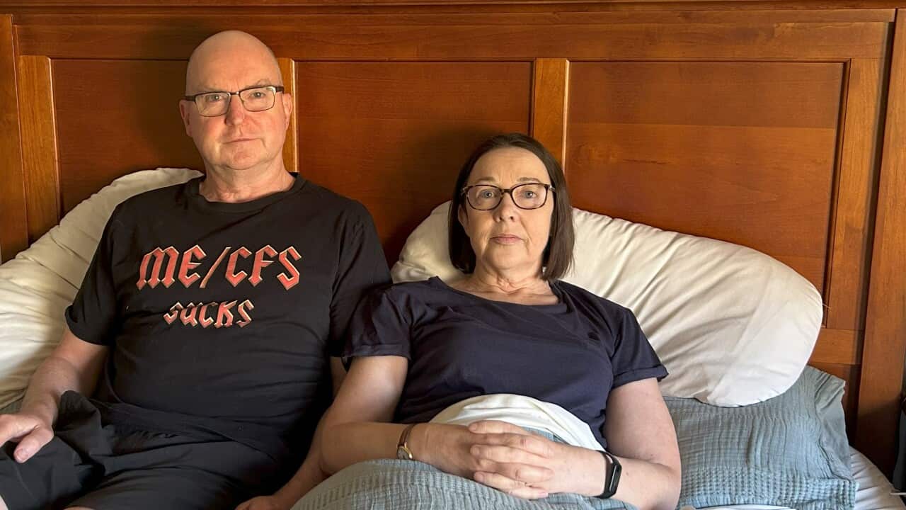 A man and a woman sit on a double bed with a neutral expression on their faces.