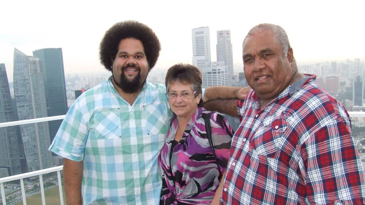 Jioji Ravulo and his parents. Jioji is wearing baggy jeans and a green, white and blue printed collared shirt. Next to him is his Mum waring a printed purple, pink and grey dress. On her other side is Jioji's Dad wearing a red printed collared shirt.