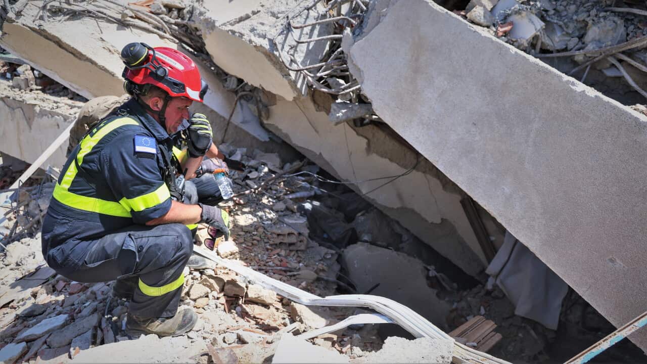 A rescue worker wearing a red helmet and high-visibility uniform crouches amidst massive, collapsed concrete rubble while communicating over a handheld radio.
