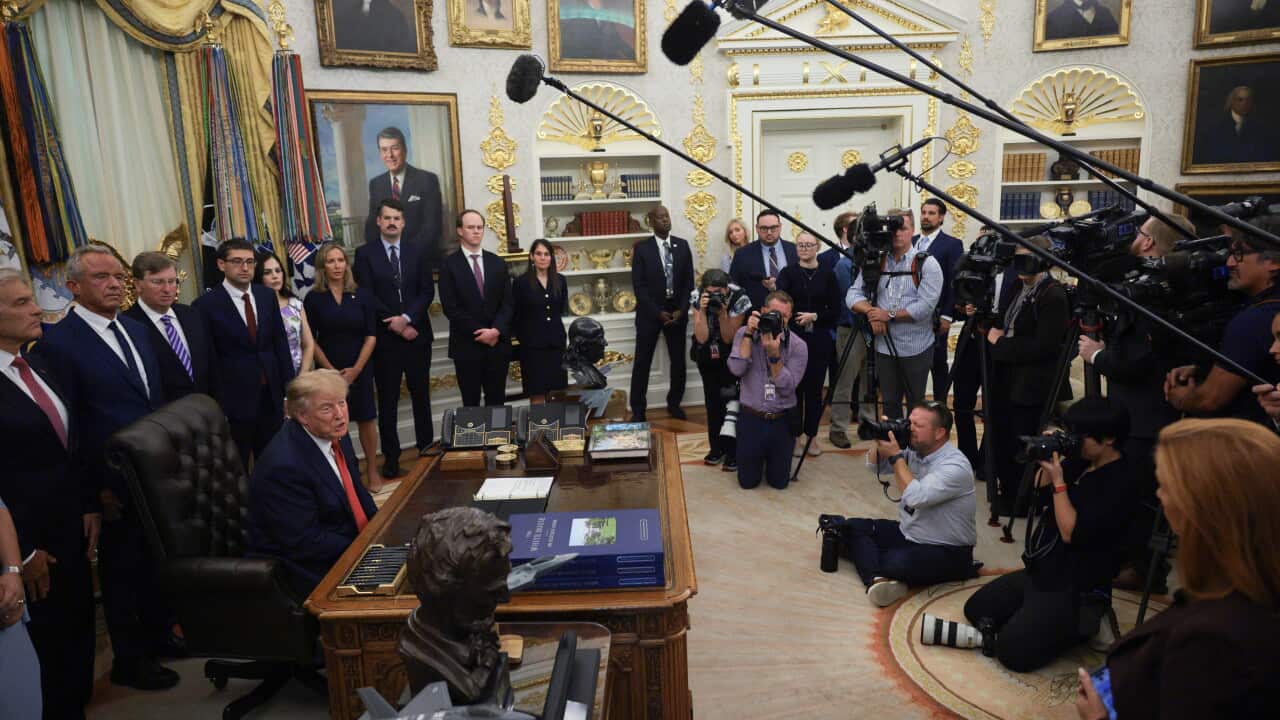 US President Donald Trump seated in the Oval Office before a large group of reporters, camera operators and photographers. Other administration officials stand behind him.