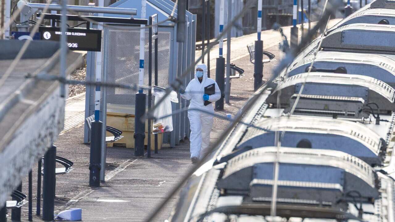 A forensic investigator wearing white protective clothing and a mask walks past a staionary train at a station.