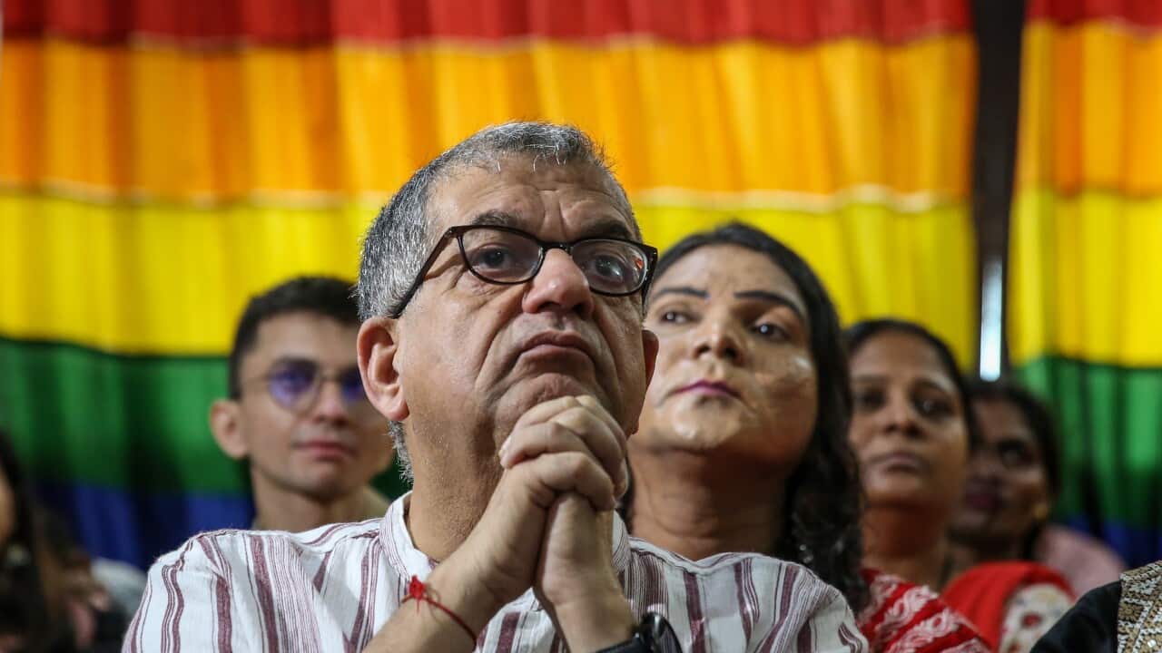 A man wearing glasses in front of a rainbow flag.