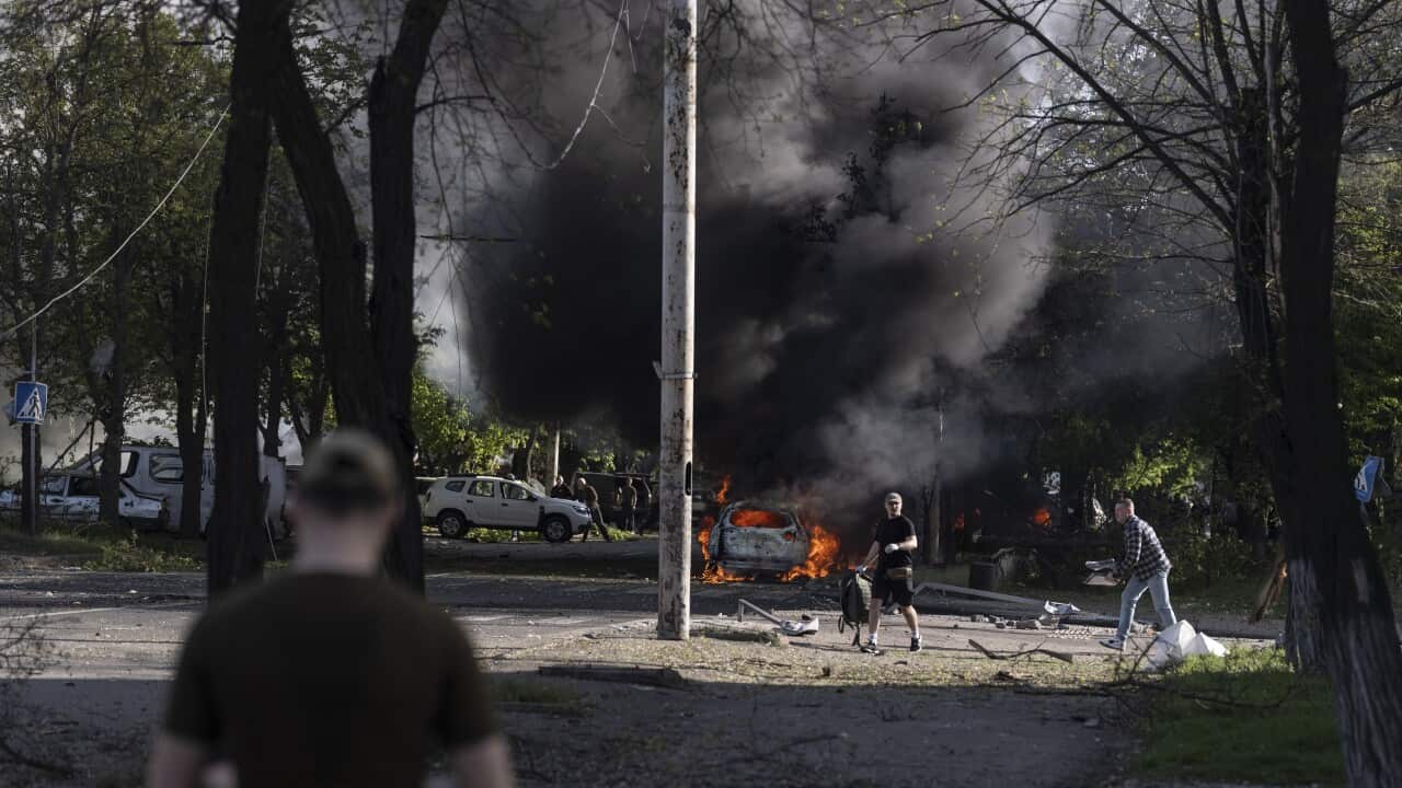 A chaotic street scene shows people walking near a vehicle engulfed in flames and a large plume of thick black smoke following an explosion.