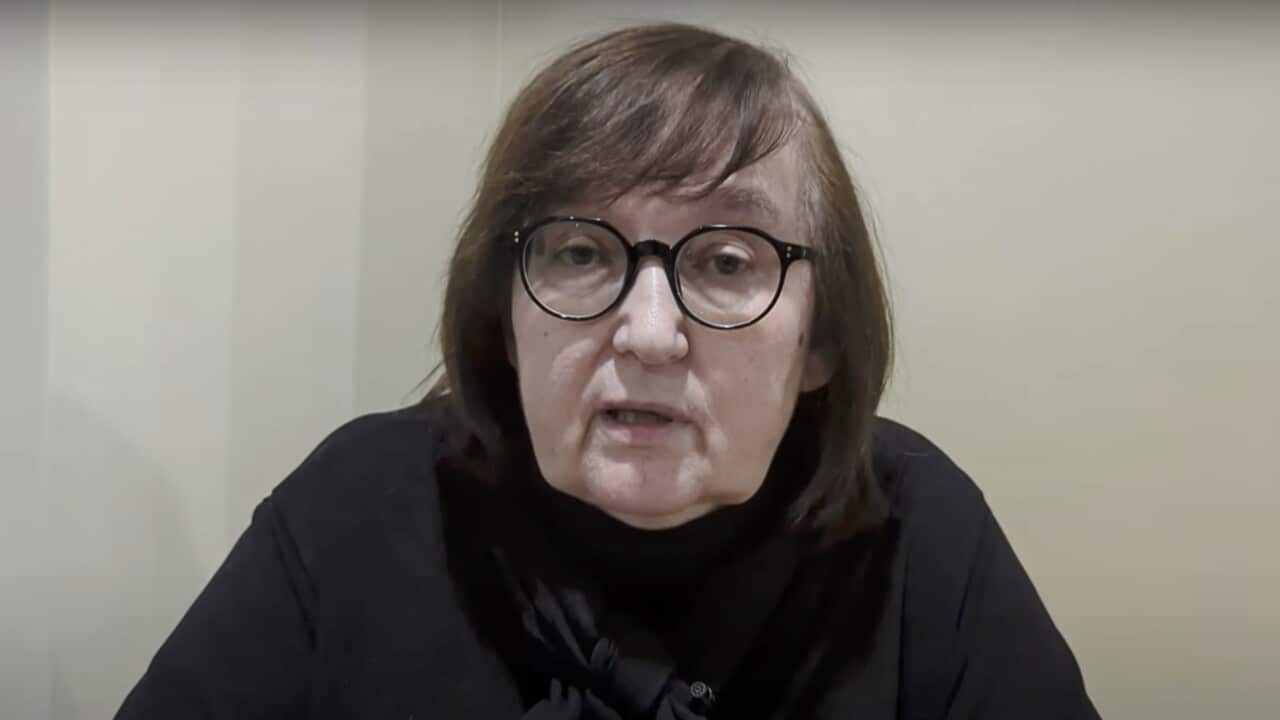 Woman in black glasses and black long sleeve top sits with top half of her body in frame.