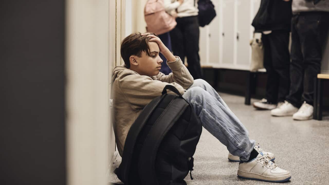 Side view of sad teenage boy sitting with backpack in school corridor