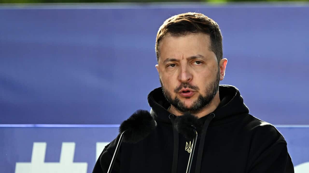 Volodymyr Zelenskyy wearing a dark top and standing behind a lectern.