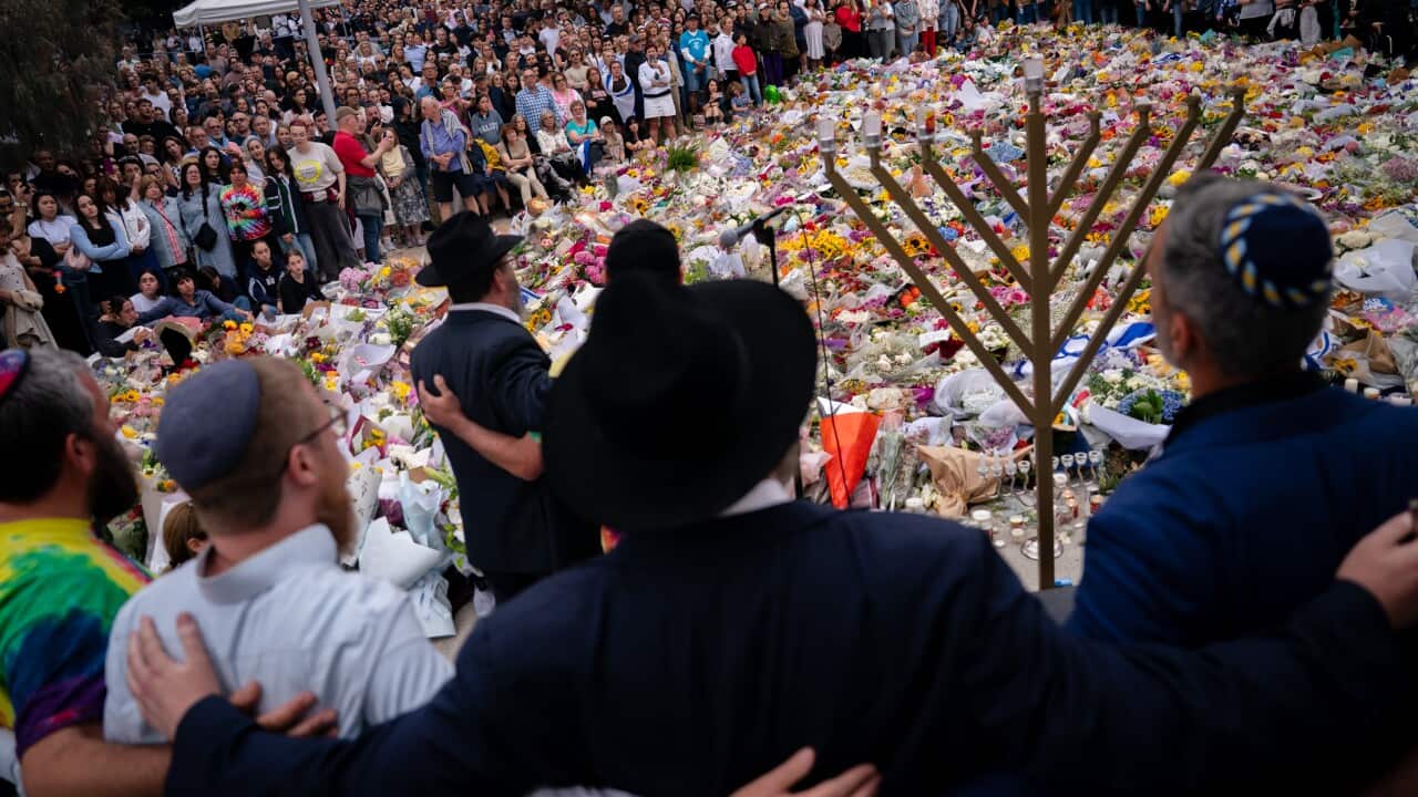 Hundreds of bouquets of flowers are surrounded by people, with a menorah and several men wearing kippahs standing with their arms around each other in the foreground.