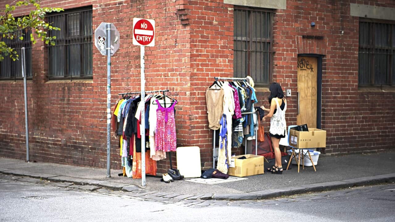 A woman looks at a rack of clothes at a garage sale on the corner of a red-brick building.