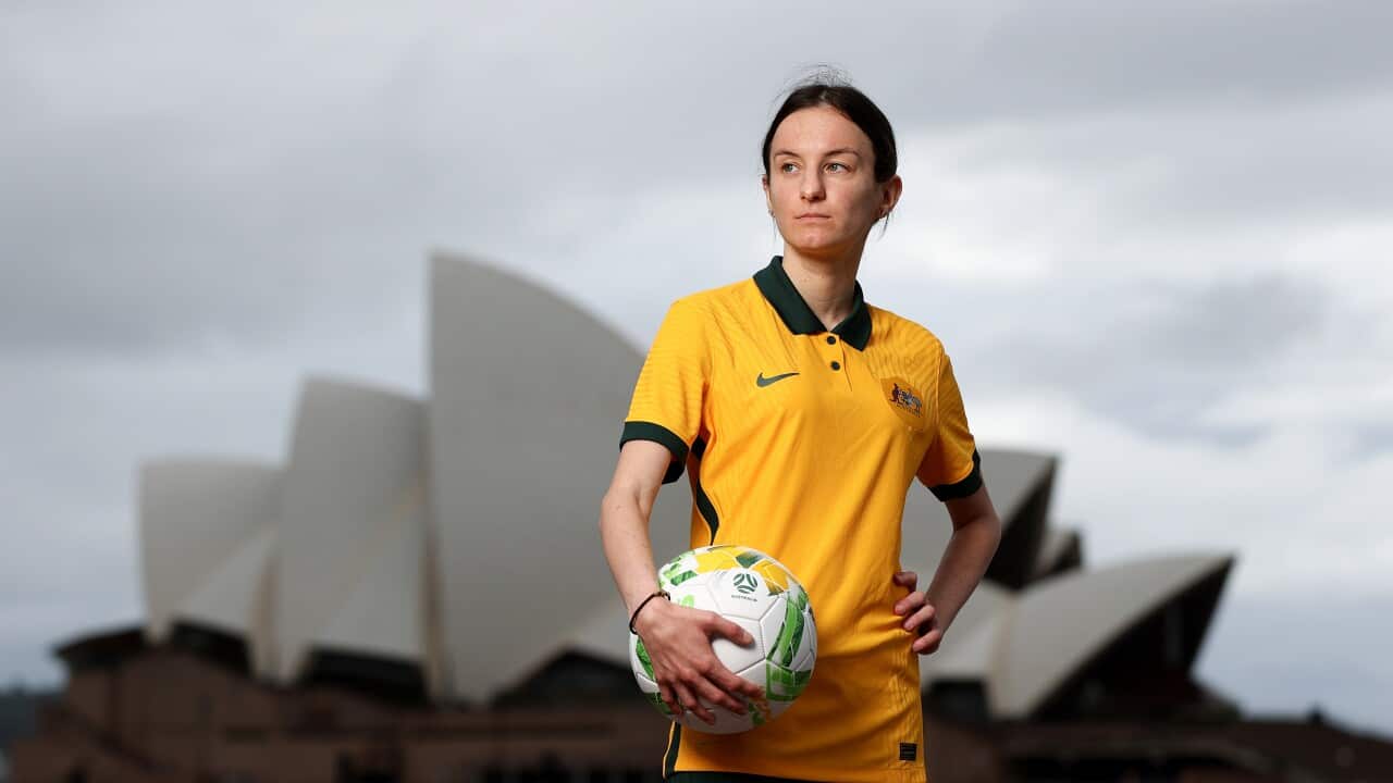 A woman in her football kit holds a soccer ball standing across the water from the Sydney Opera House.