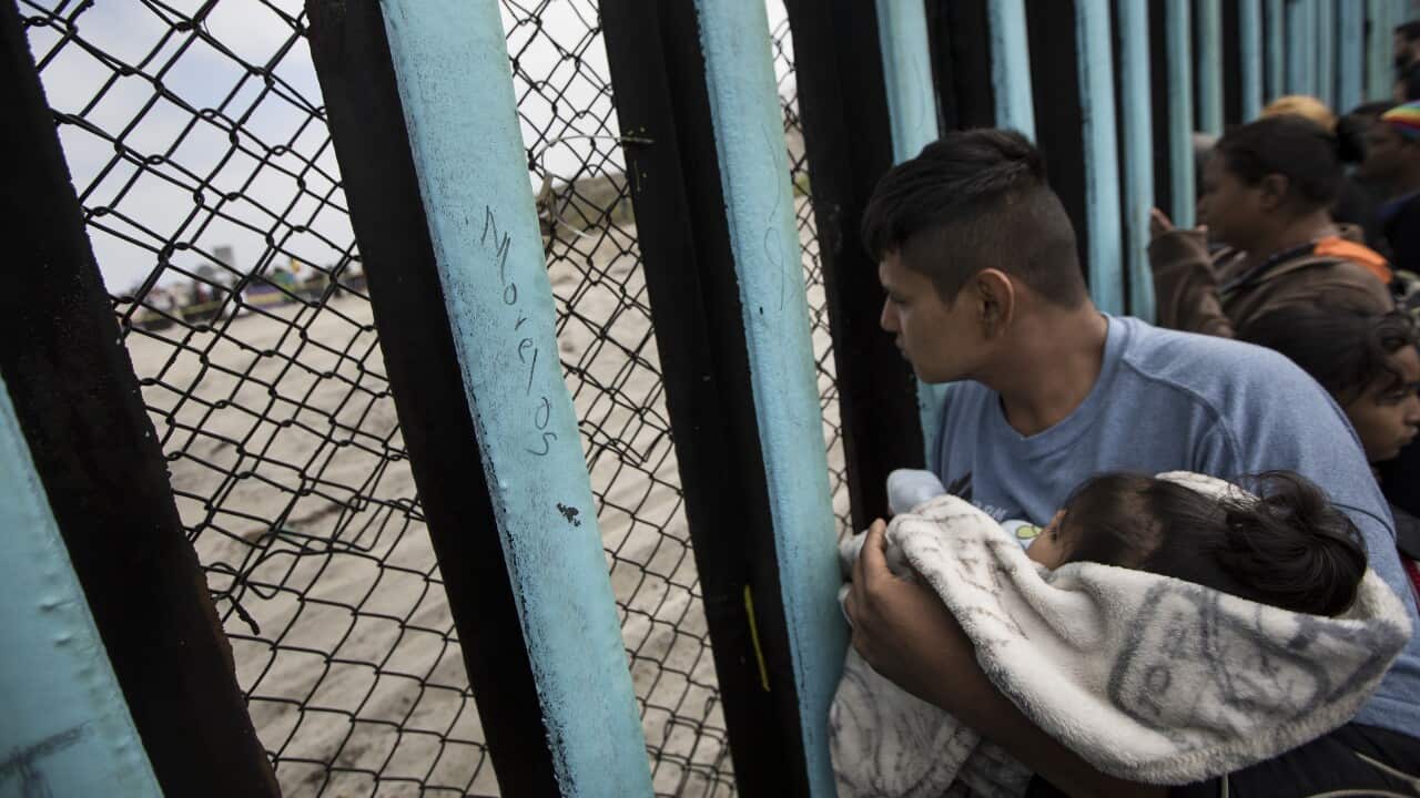 A member of the Central American migrant caravan, holding a child, looks through the border wall