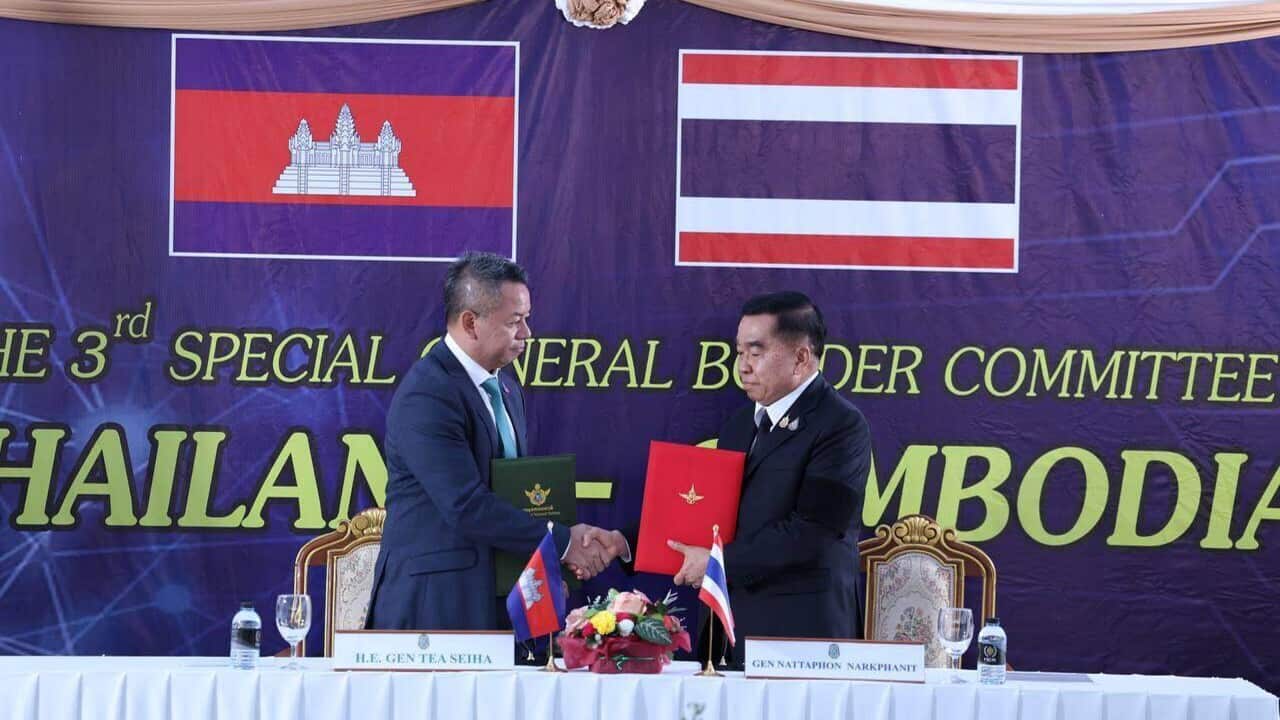 Two officials wearing suits shake hands while holding folders in front of the flags of Thailand and Cambodia.