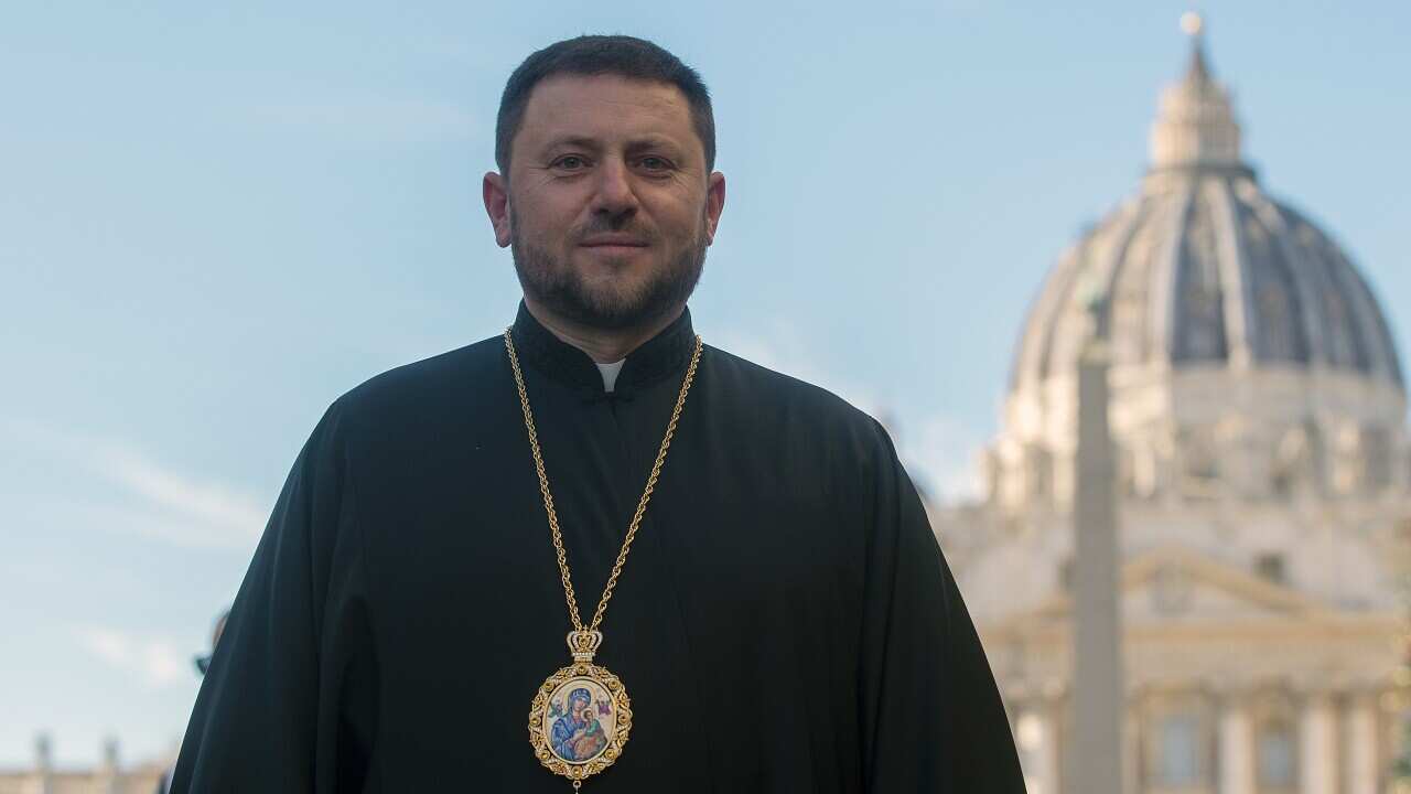 A man in a black religious robe with a large circular locket worn around his neck on a long chain is posing for a picture.