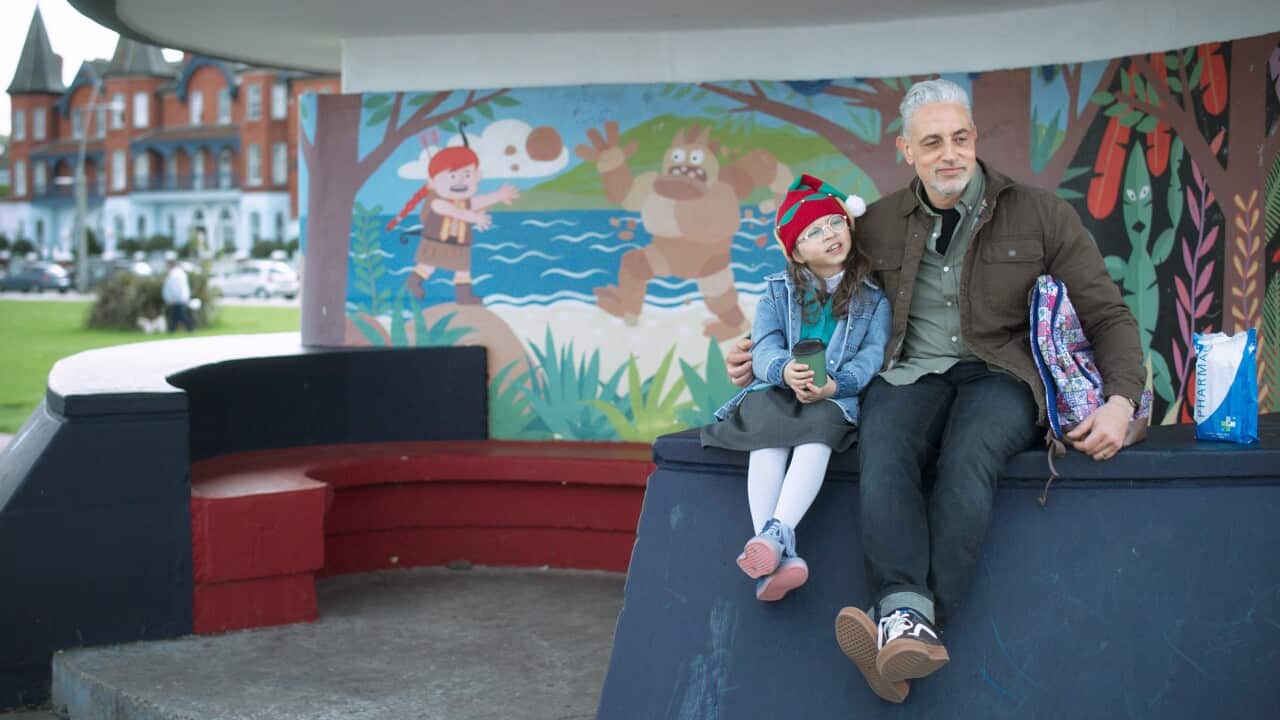 A man and a young girl sit on a stone bench in front of a bright mural. He has his arm around her shoulder.