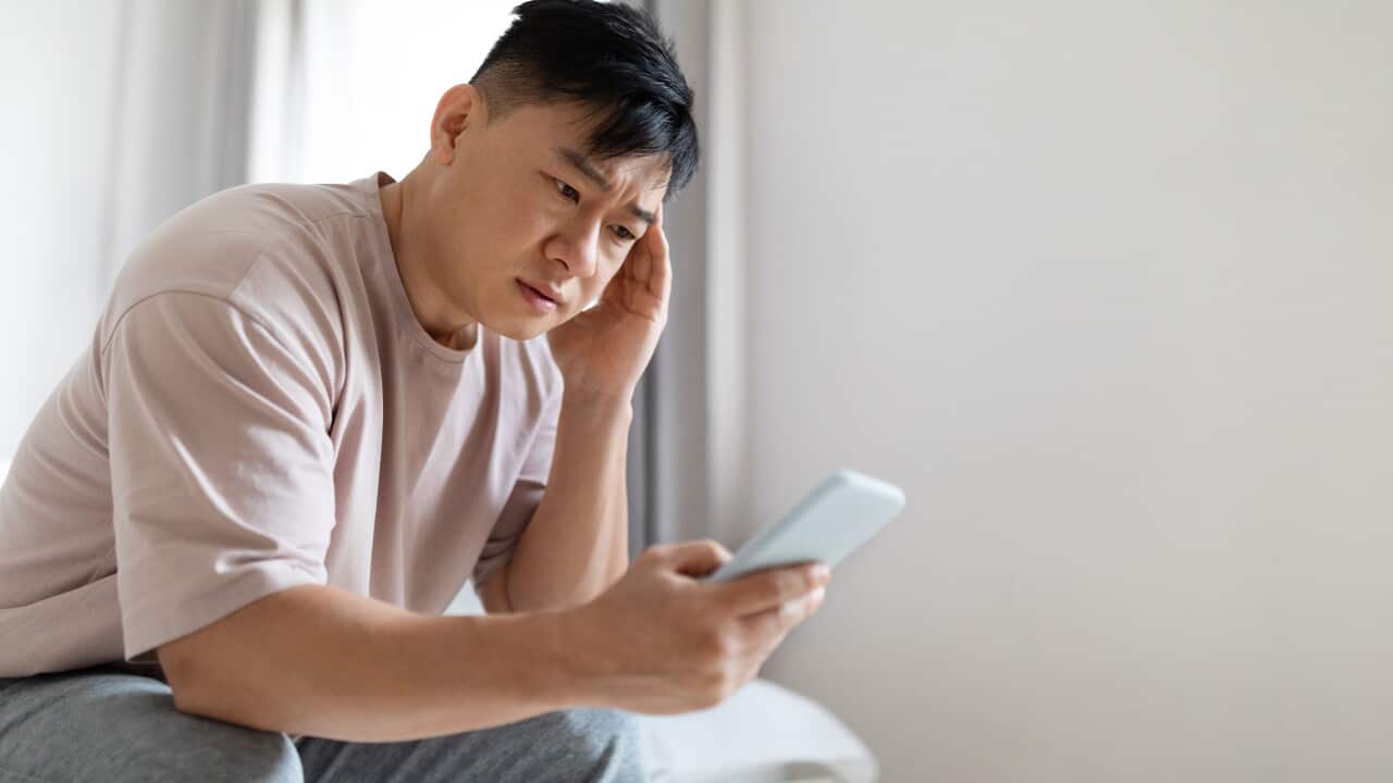 Worried upset asian man sitting on bed, using smartphone