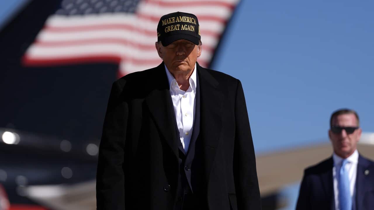 A man wearing a black trenchcoat and a cap walking in front of an aeroplane