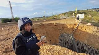 A woman wearing a press vest and helmet reports from a destroyed site