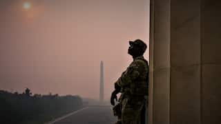 A solider leans against a column with the Washington Monument behind him and a red sun in the sky as smoke obscures much of the background.